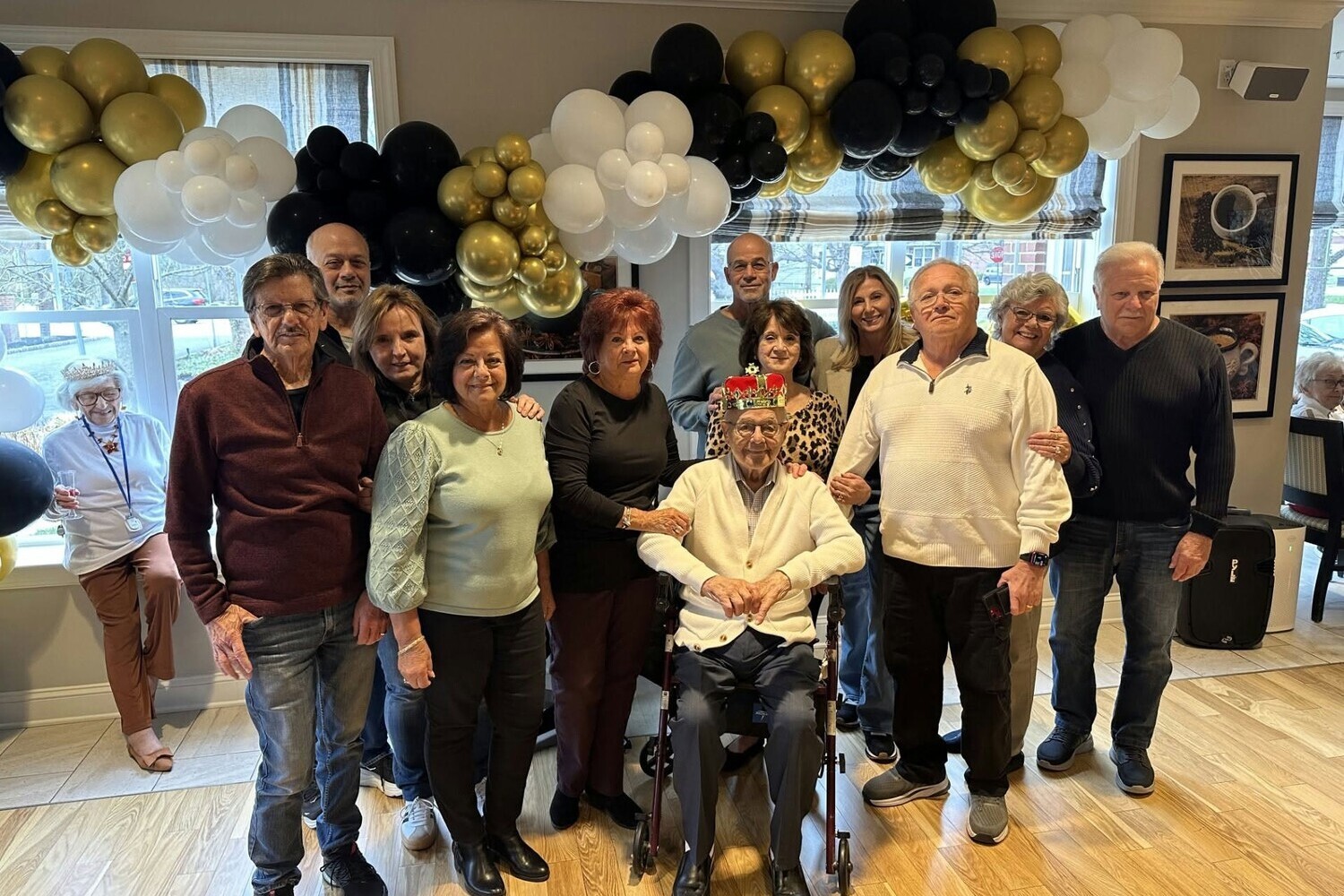 Residents, family, and staff celebrate together indoors with festive black, gold, and white balloon decor, capturing a joyful special event at a senior living community in Morris Plains, NJ.
