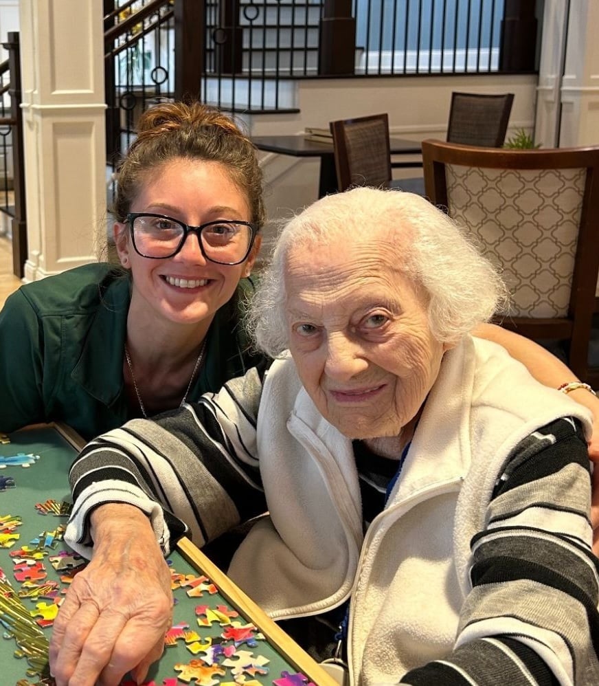 A staff member and a resident share a smile while working on a colorful puzzle together in a bright common area at Arbor Terrace Norwood, a senior living community in Norwood, NJ.