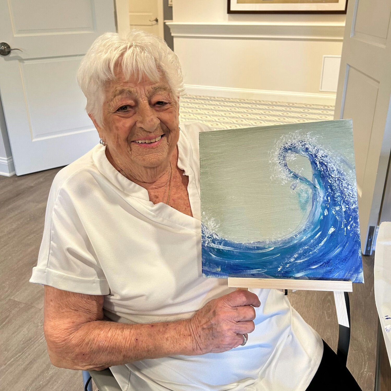 A resident at Arbor Terrace Norwood smiles while holding a wave-themed painting, celebrating creativity at this senior living community in Norwood, NJ.