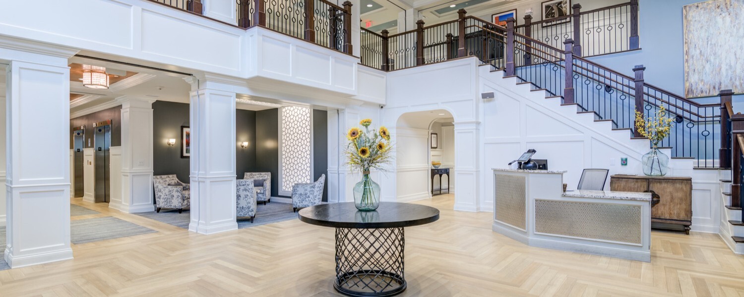 Bright, two-story lobby at Arbor Terrace Norwood featuring a curved staircase, herringbone wood floors, a round table with fresh sunflowers, and cozy seating, part of a senior living community in Norwood, NJ near Bergen County.