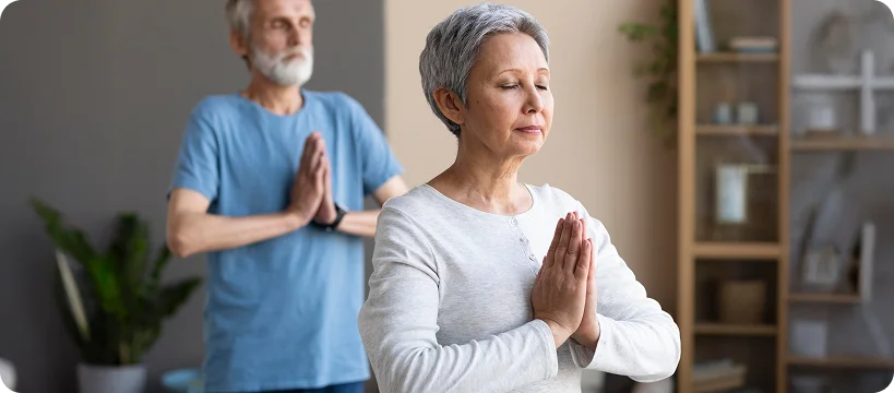 A senior woman and man practice yoga together with their hands in a prayer pose.
