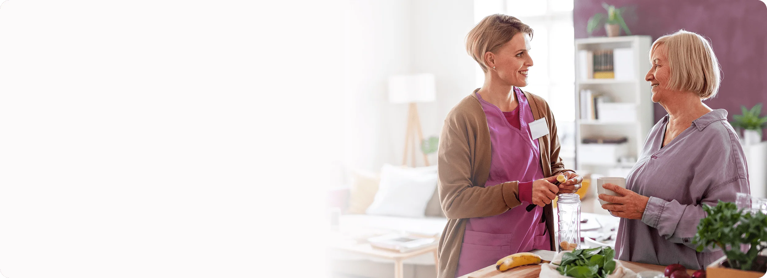 A smiling female caregiver holds a snack while talking to a happy senior woman holding a mug.