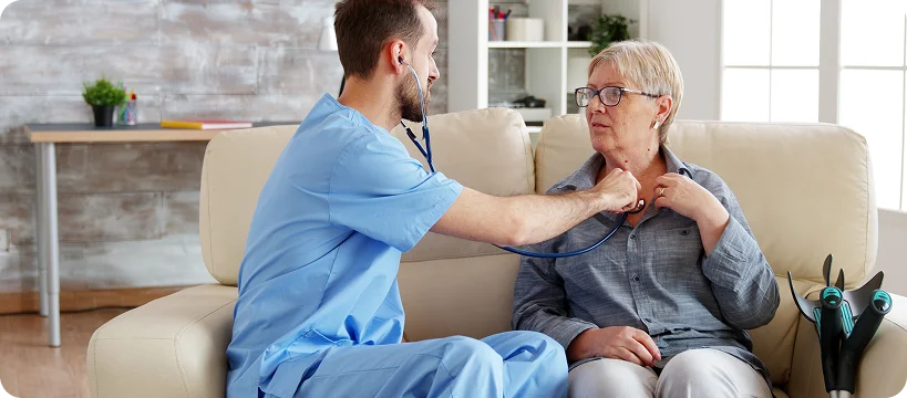 A male nurse or caregiver listens to an elderly woman’s heartbeat with a stethoscope while she sits on a couch.