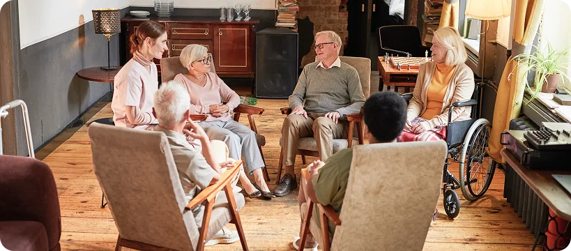 A small group of seniors and a female caregiver sit in a circle in comfortable chairs, engaged in a conversation.