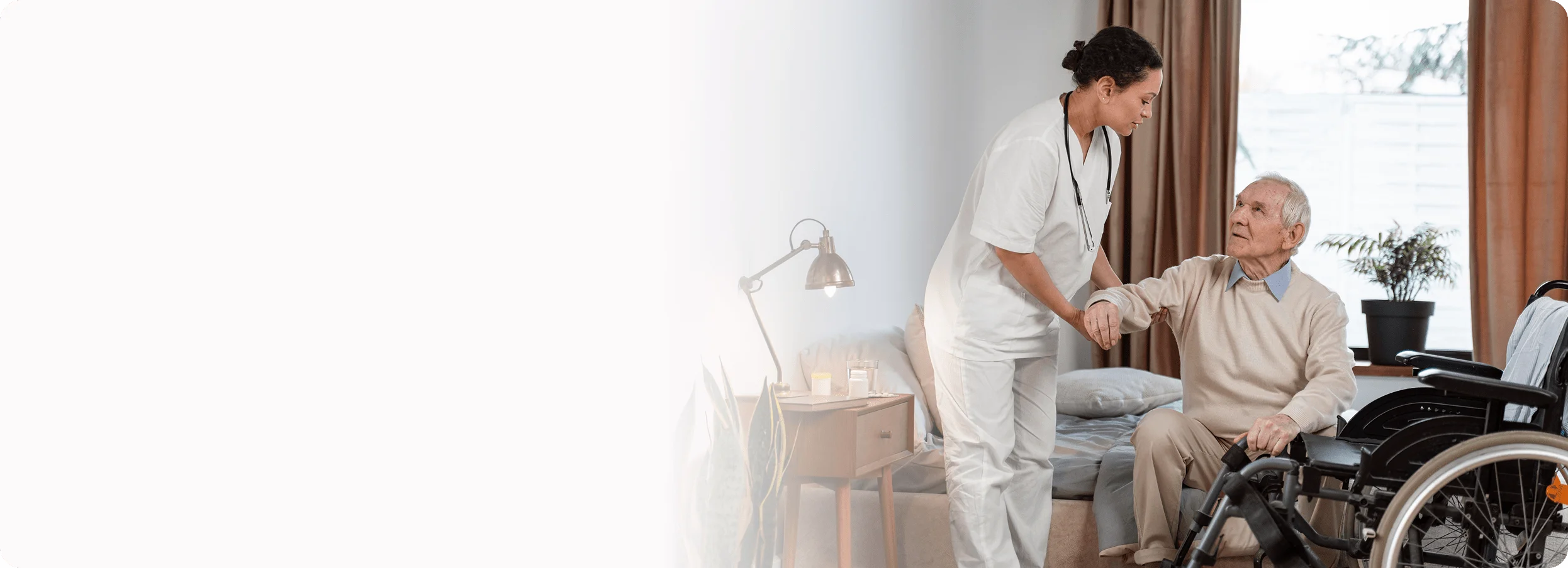 A female caregiver in a white uniform helps an elderly man stand up from his chair.