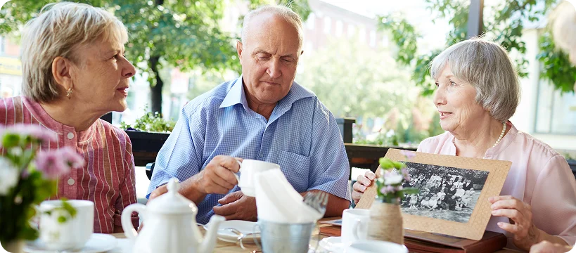 An elderly man and two elderly women sit at an outdoor cafe table, looking at an old framed black-and-white family photo.