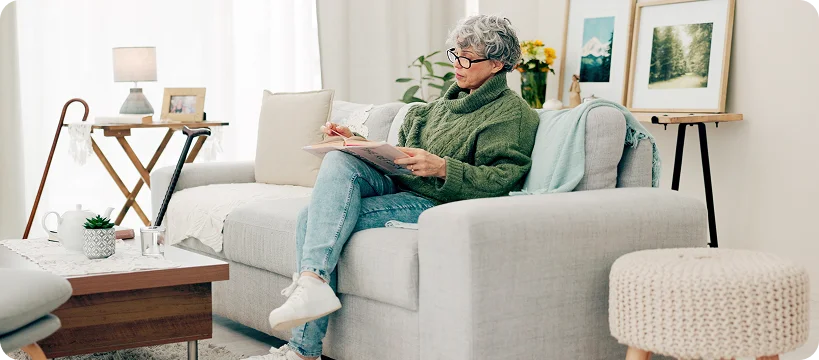 A senior woman wearing glasses sits on a couch in a living room, reading a book.