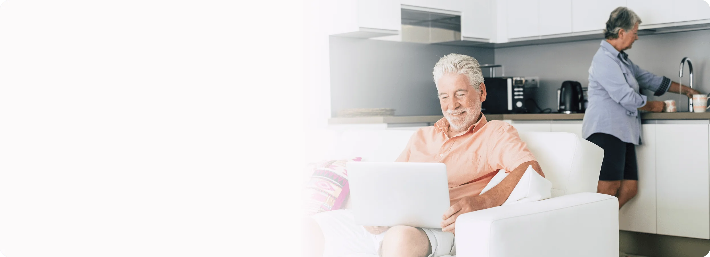 An elderly man smiles while using a laptop on a couch as an elderly woman washes dishes in the background.