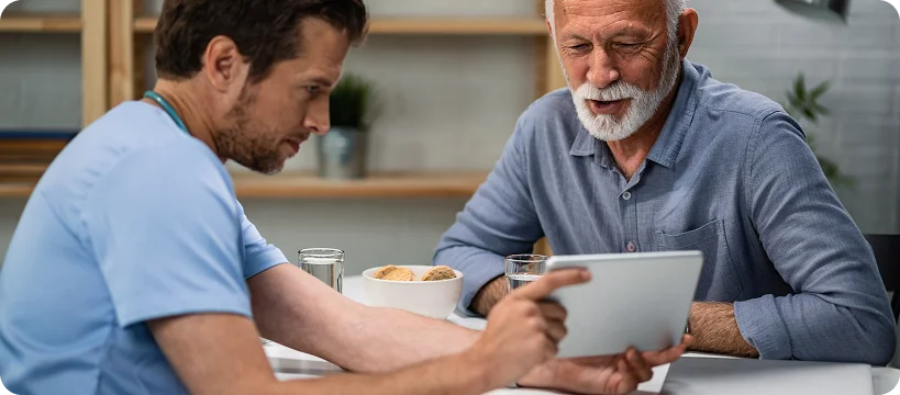 A young male caregiver sits with an elderly man at a table, showing him something on a tablet.