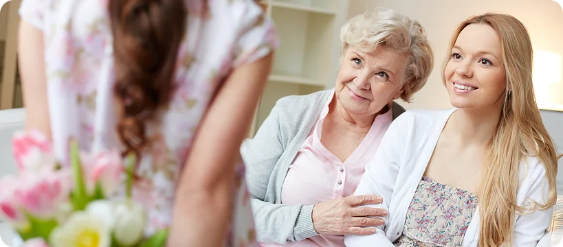 An elderly woman and a younger woman smile as a child approaches with a bouquet of flowers.
