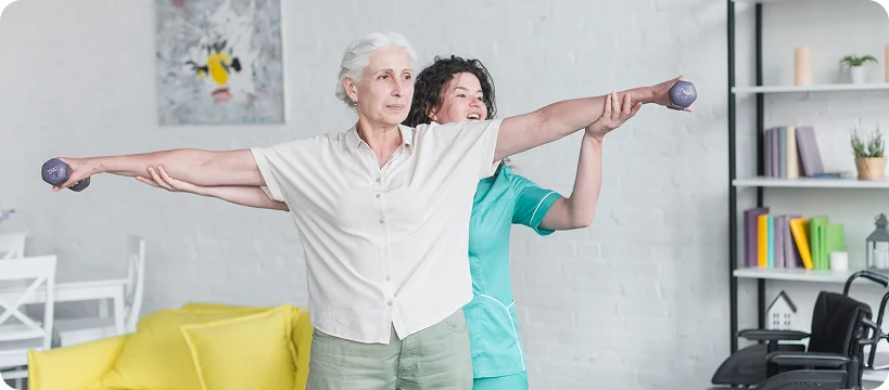 A physical therapist helps an elderly woman with her arm exercises, holding her arm and a small dumbbell.