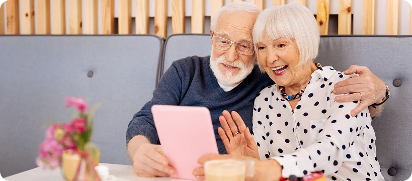 A joyful elderly couple smiles and waves at a screen during a video call on a tablet.